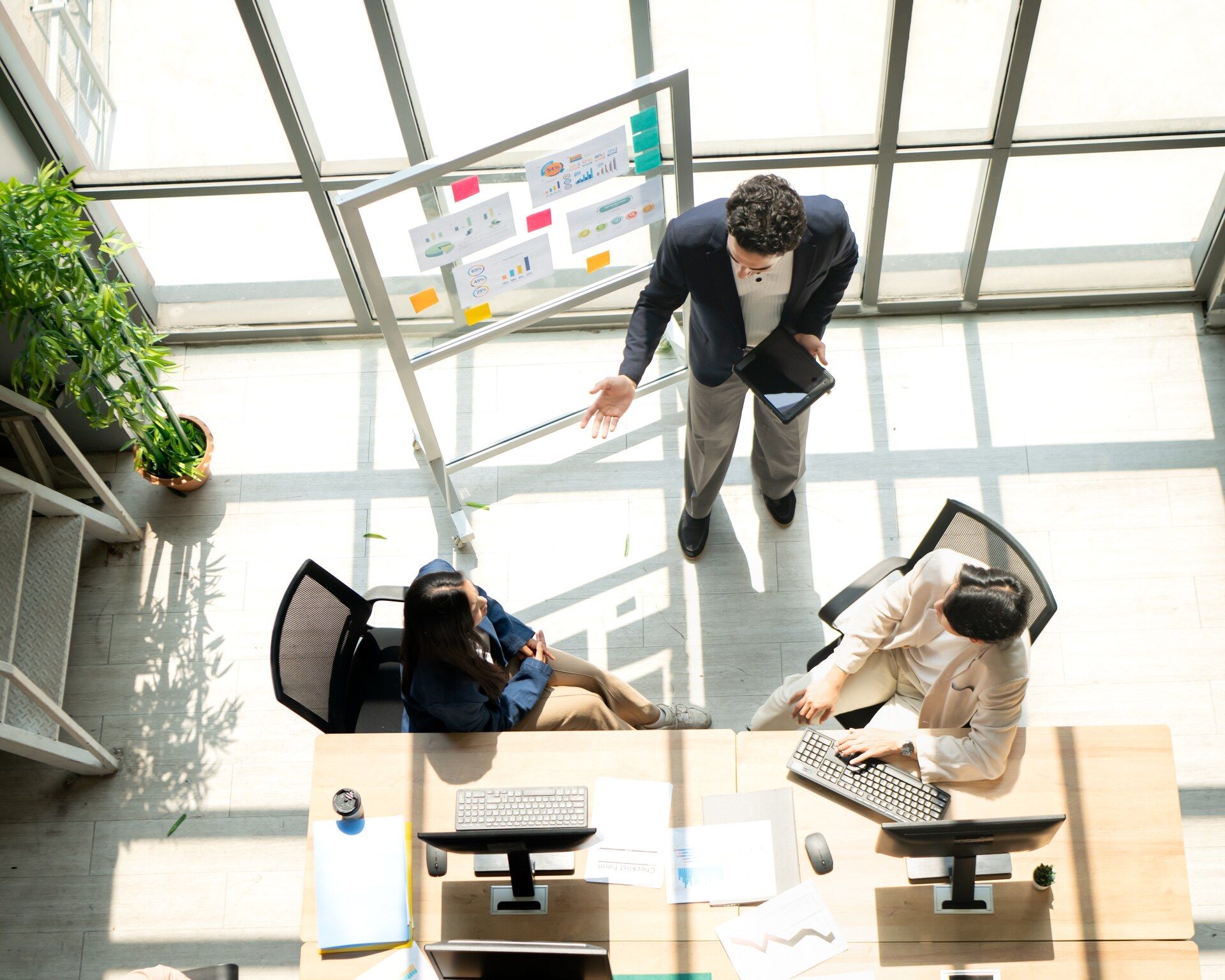 A team seen from above, gathered around a shared desk as a colleague presents charts in a glass-walled office